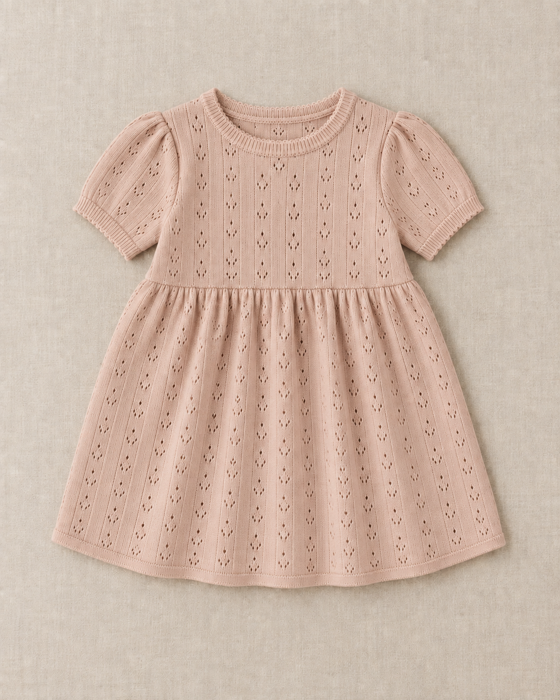 Blush pointelle knit baby dress with short puff sleeves, a ribbed crew neck and a gathered skirt, laid flat on a linen surface.
