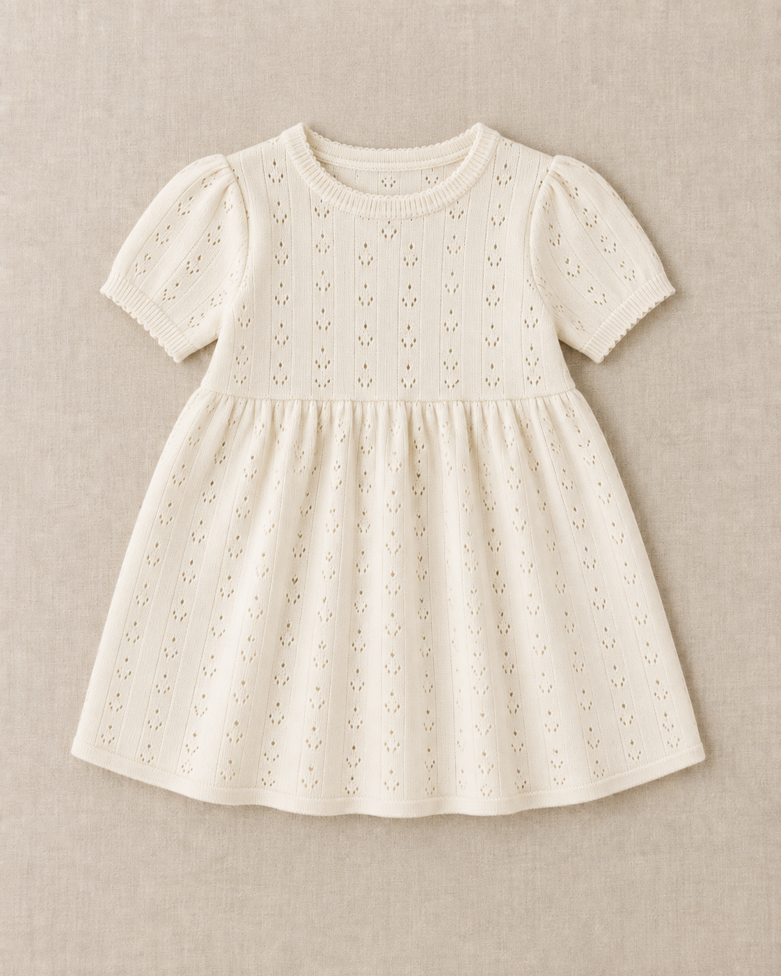 Cream pointelle knit baby dress with short puff sleeves, a ribbed crew neck and a gathered skirt, laid flat on a linen surface.