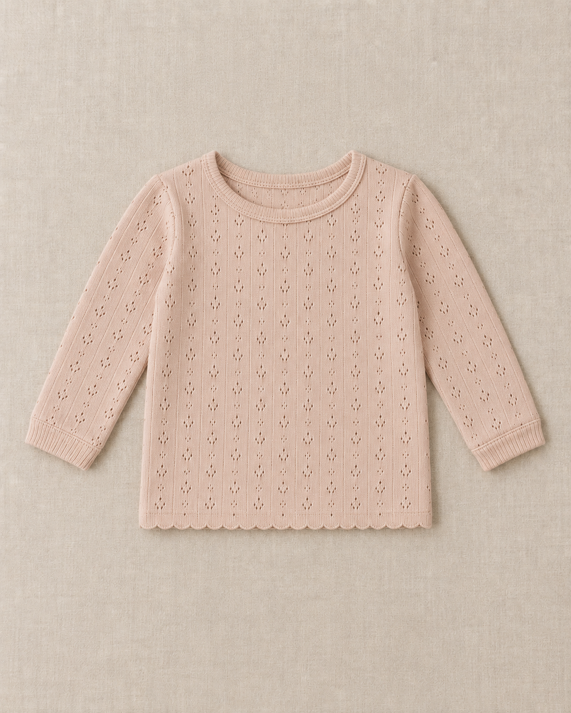 Blush pointelle knit long sleeve top with a ribbed crew neck, ribbed cuffs and a straight scalloped hem, laid flat on a linen surface.