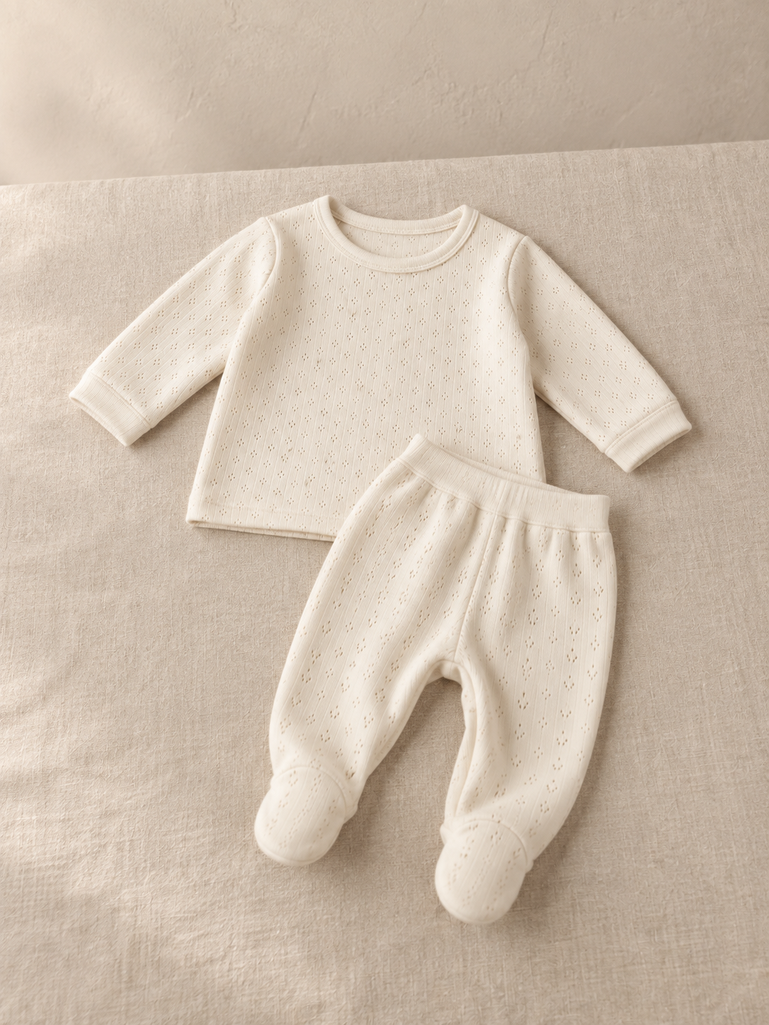 Cream pointelle knit two-piece set laid out on a linen surface, the long sleeve top above and the matching footed bottoms below.