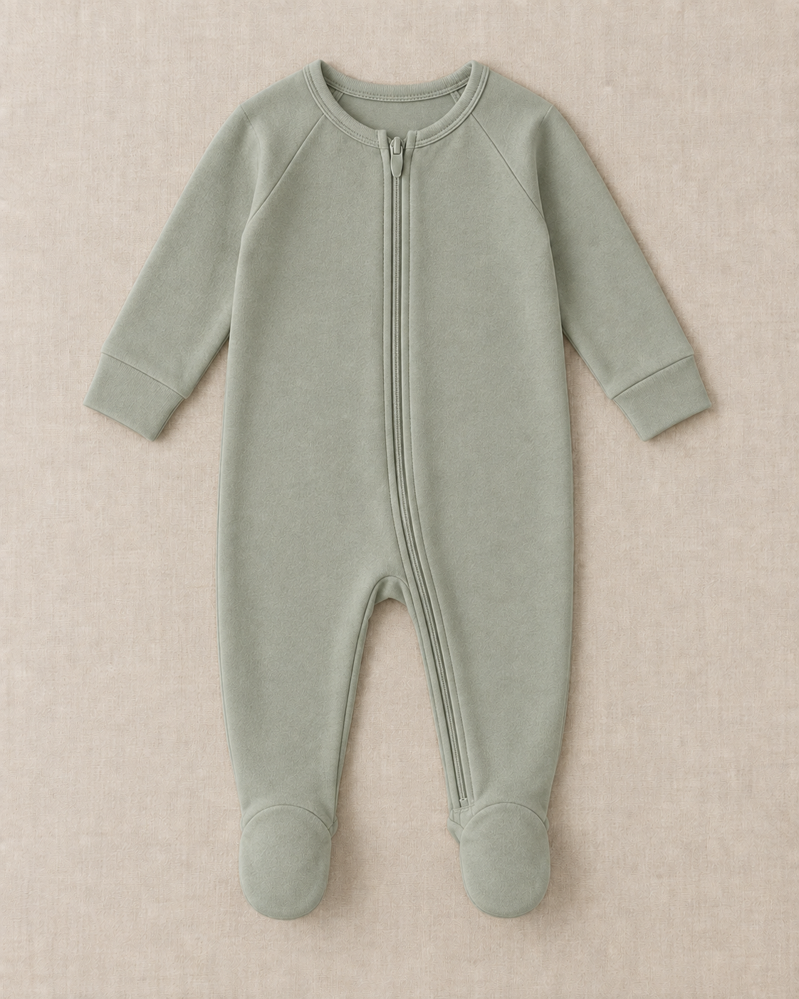 Sage footed sleep suit with long raglan sleeves and a full-length front zip, laid flat on a linen surface.
